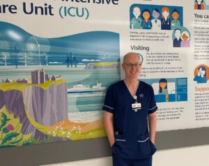 Steve Marjoribanks standing in front of a visitor information sign at Aberdeen Royal Infirmary's Critical Care Unit. The sign is white and shows an illustrated image of Dunottar Castle with the sea in the background. Some text is visible on the sign in the background.