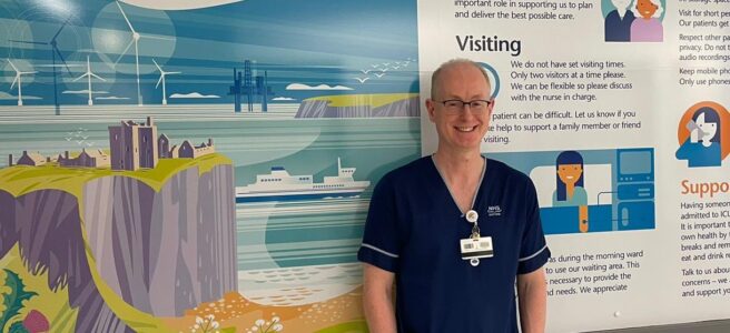 Steve Marjoribanks standing in front of a visitor information sign at Aberdeen Royal Infirmary's Critical Care Unit. The sign is white and shows an illustrated image of Dunottar Castle with the sea in the background. Some text is visible on the sign in the background.