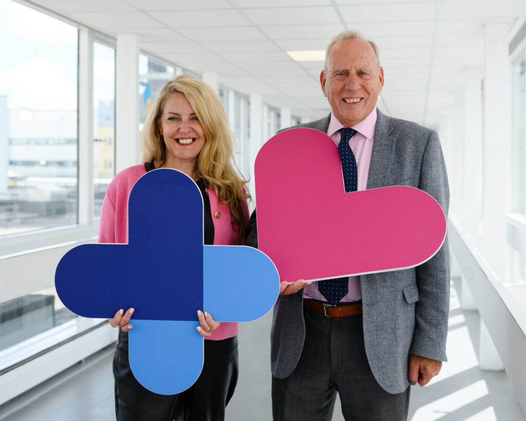 Image shows Lisa Duthie. She has blonde, shoulder-length hair and is wearing a pink blazer and black trousers. She is holding a large image of the plus sign from the charity logo, which is dark blue at the top and light blue at the bottom. Standing beside her is Dennis Robertson. He is wearing a grey suit with a white shirt and red tie. He is holding a large image of the pink heart from the charity logo. They are standing in a long hospital corridor which has grey flooring and light walls.