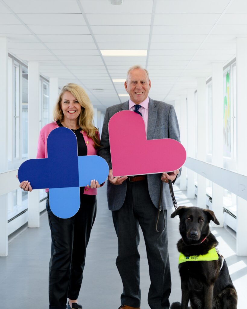 Image shows Lisa Duthie. She has blonde, shoulder-length hair and is wearing a pink blazer and black trousers. She is holding a large image of the plus sign from the charity logo, which is dark blue at the top and light blue at the bottom. Standing beside her is Dennis Robertson. He is wearing a grey suit with a white shirt and red tie. He is holding a large image of the pink heart from the charity logo. He is holding a lead, and his guide dog is sitting by his feet. The dog is black and is wearing a yellow vest. The three are standing in a long hospital corridor which has grey flooring and light walls.