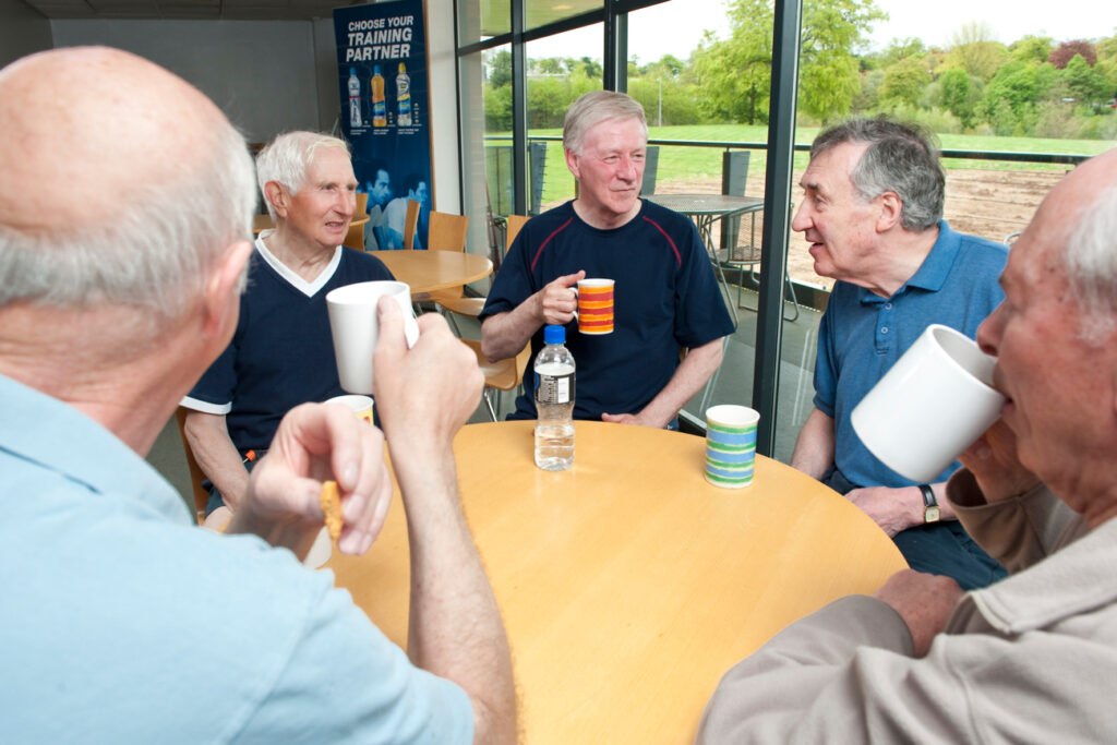 Image shows a group of five men sitting around a circular table. They are drinking from mugs of tea and conversing with each other.
