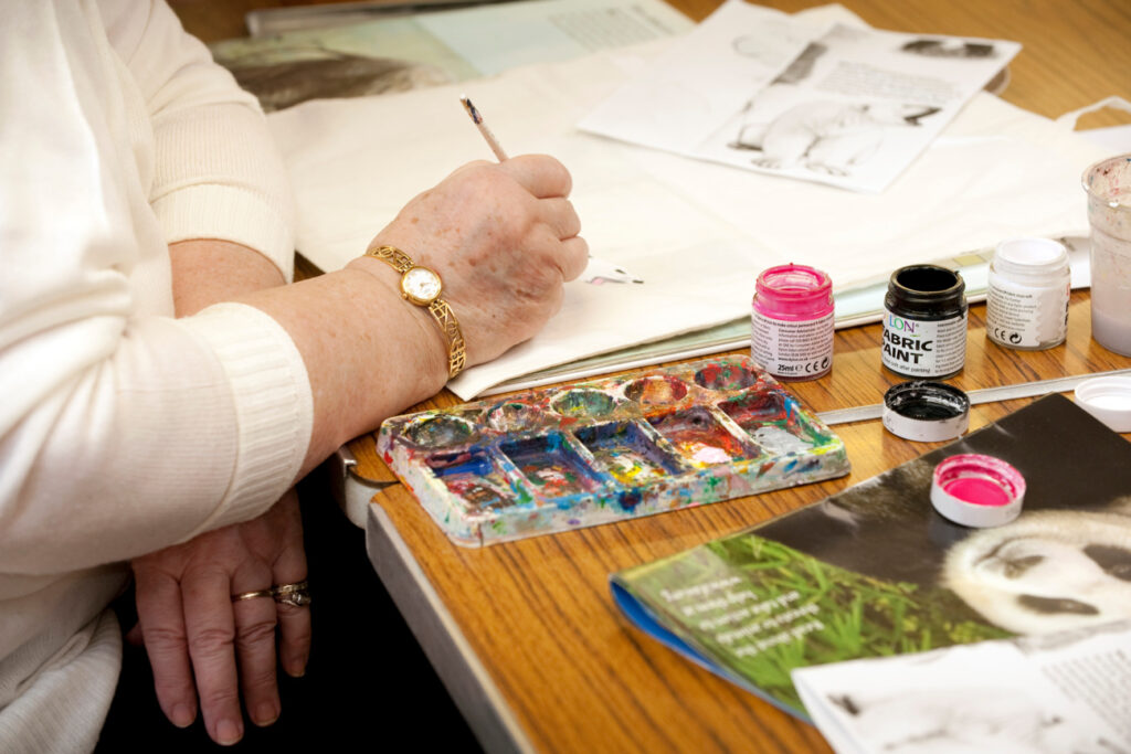 Image shows a close up of a lady painting. She is wearing a white jumper, with only her hands and arms visible. Art supplies including pots of pink, black and white paint and a mixing palette are arranged beside her. She is painting a polar bear with its mouth open, using a printed image for reference.