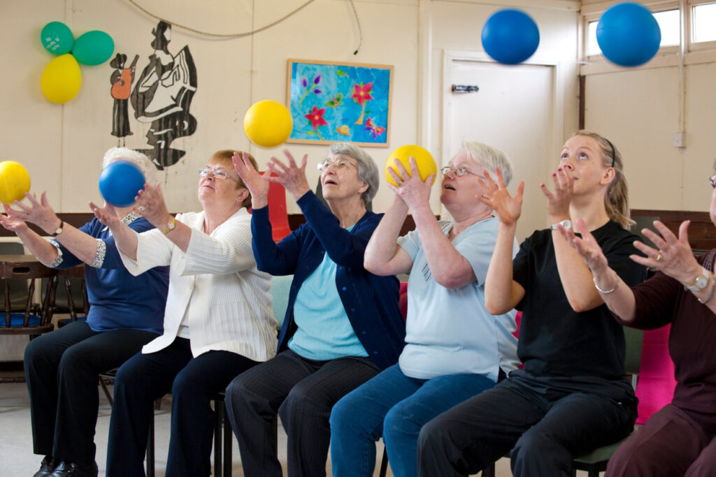 The image shows five women sitting on a row of plastic chairs. The woman to the right of the photo has blonde hair tied in a ponytail and wears a black t-shirt with black trousers. The other women have grey hair and wear glasses. They are wearing a mixture of t-shirts, jumpers and cardigans. All the women have thrown small yellow and blue yoga balls into the air and are looking at the balls as they try to catch them.