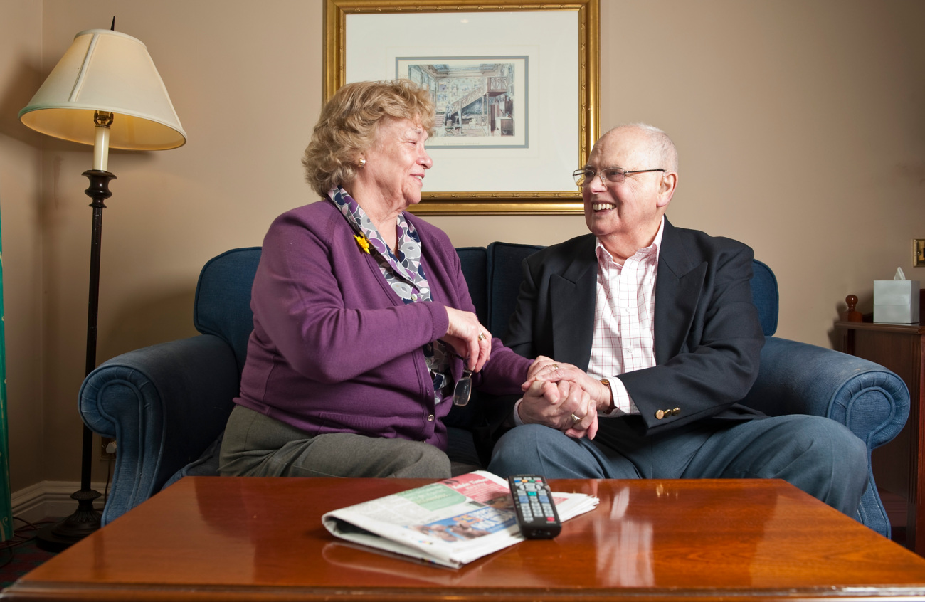 The image shows an older couple sitting on a sofa in a living room. They are holding hands and smiling at each other. The lady has short, curled hair and wears a purple garden with a purple and grey floral blouse. The man wears a pink checked shirt with a black blazer.