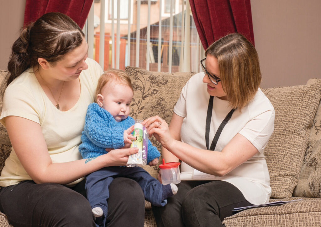 Image shows a mum with a baby on her lap, talking to another lady. The mum is wearing a pale yellow t-shirt and has long, brown hair. She is holding a small baby, who is wearing a blue knitted jumper. The other lady is wearing a white t-shirt and has short brown hair. She is holding the baby's sip cup, and handing the mum and baby a cellophane-wrapped toothbrush and toothpaste.