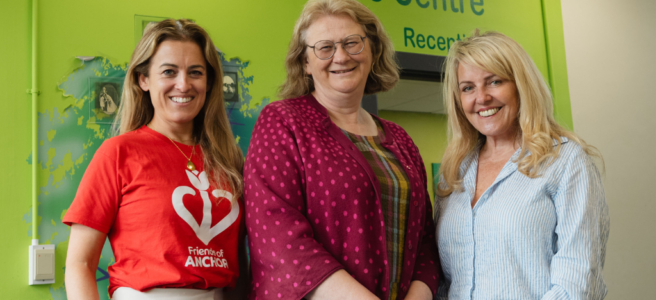 The image shows three women standing in front of a green-painted wall which was 'Welcome to Clinical Genetics Centre' written on it. The lady on the left wears a red t-shirt featuring the Friends of ANCHOR logo and white trousers. She has long brown hair, and stands with one hand in her pocket. The lady in the middle has chin-length brown hair and glasses. She wears checked dress and a dark pink cardigan with lighter polka dots. The lady on the right has blonde hair and wears a light blue shirt and black trousers. She stands with her hands clasped in front of her. All three women are looking at the camera and smiling.