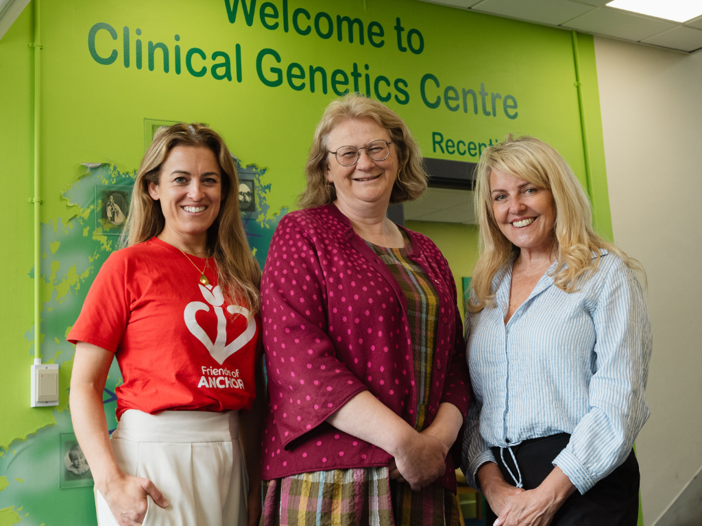 The image shows three women standing in front of a green-painted wall which was 'Welcome to Clinical Genetics Centre' written on it. The lady on the left wears a red t-shirt featuring the Friends of ANCHOR logo and white trousers. She has long brown hair, and stands with one hand in her pocket. The lady in the middle has chin-length brown hair and glasses. She wears checked dress and a dark pink cardigan with lighter polka dots. The lady on the right has blonde hair and wears a light blue shirt and black trousers. She stands with her hands clasped in front of her. All three women are looking at the camera and smiling.